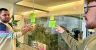 Two colleagues pointing at sticky notes on a glass wall, outlining a consolidated framework for managing product information.