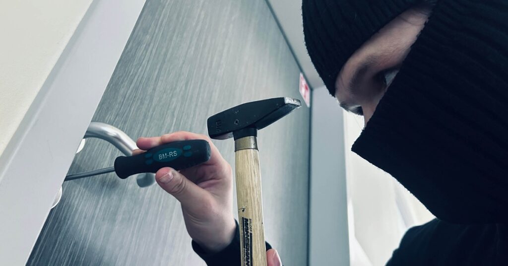 Person wearing dark clothing trying to break open a door lock with a screwdriver and hammer.