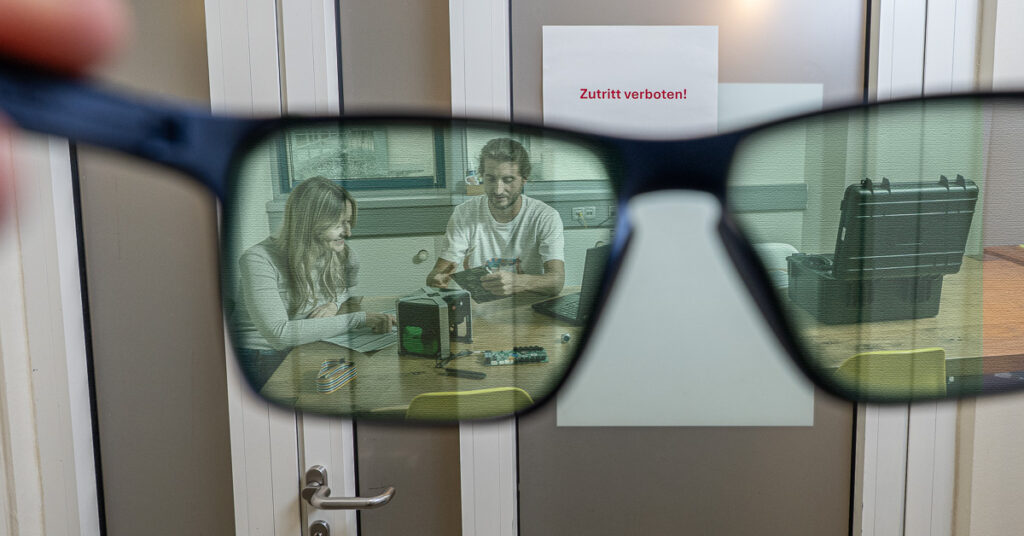 A pair of glasses held up against a closed door offers a view of two people working on a product at a table.
