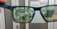 A pair of glasses held up against a closed door offers a view of two people working on a product at a table.
