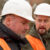 Close-up of two people wearing protective helmets and orange safety vests at a construction site.